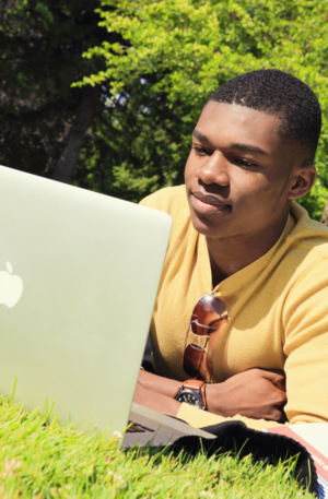 Male student works on his laptop while laying on the grass.